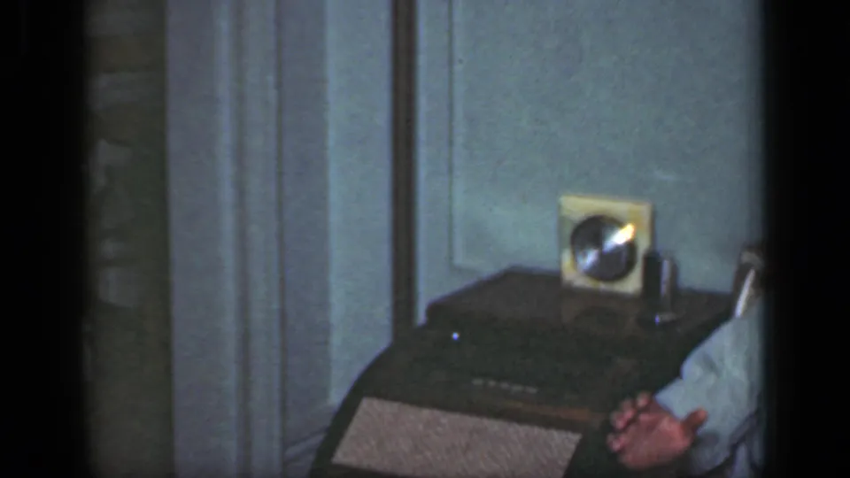 A 1949 New York living-room frame shows a radio and clock on a table with a seated person's hands visible at the sofa edge.