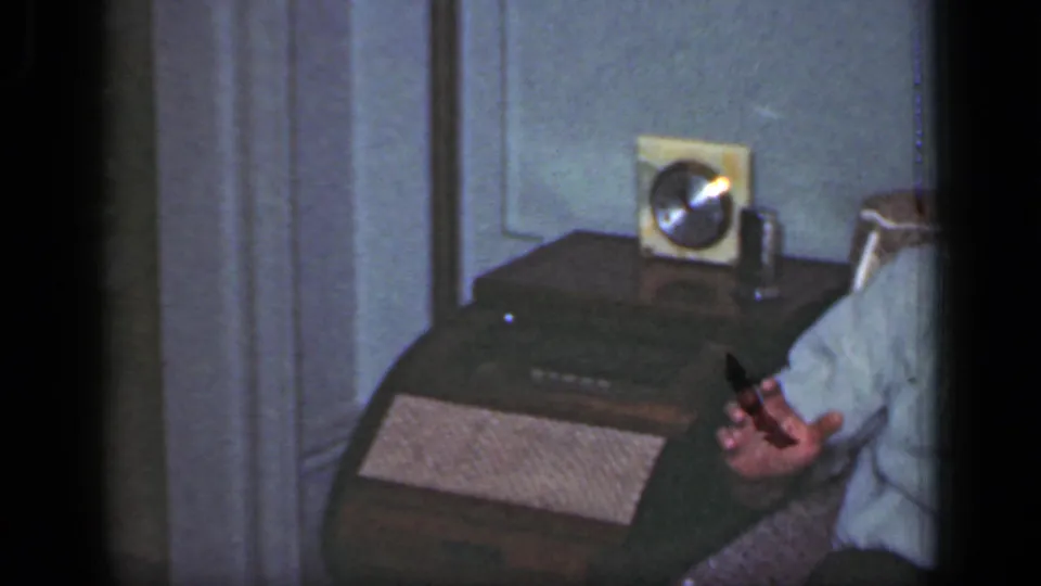 1949 New York home-movie frame showing part of a man in an armchair beside a radio on a wooden side table.