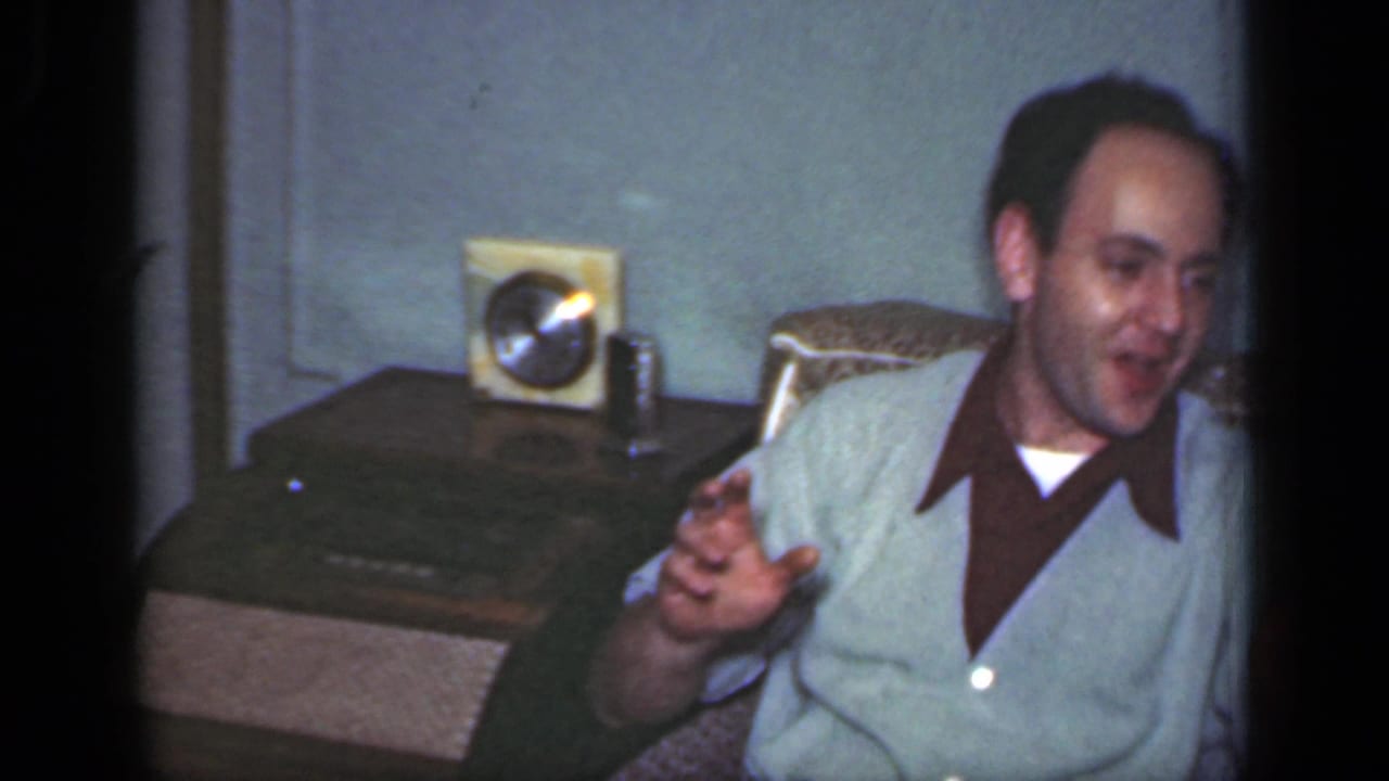 1949 New York frame of a smiling man in a light jacket gesturing from a patterned armchair in a private room.