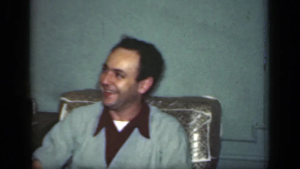 1949 New York frame showing a seated man smiling to the side in a dim private living-room setting.