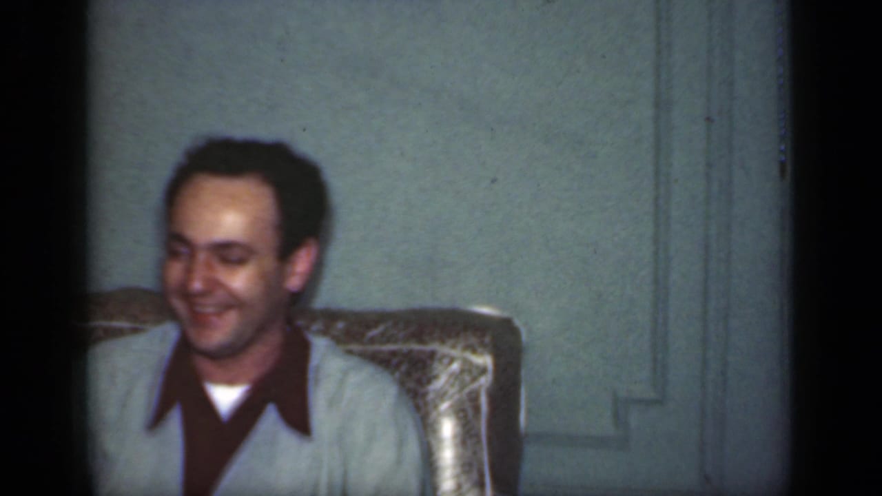 A smiling man sits in a patterned armchair against a pale wall.
