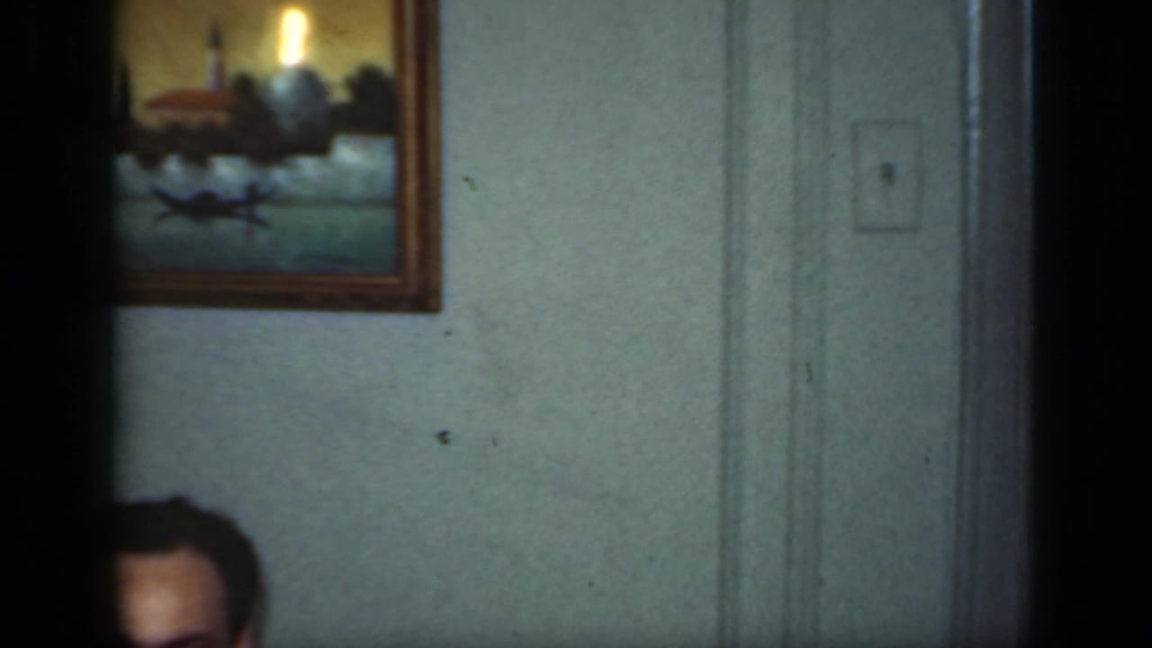The camera drifts across the home interior, leaving only the top of the man's head visible beneath a framed coastal painting.