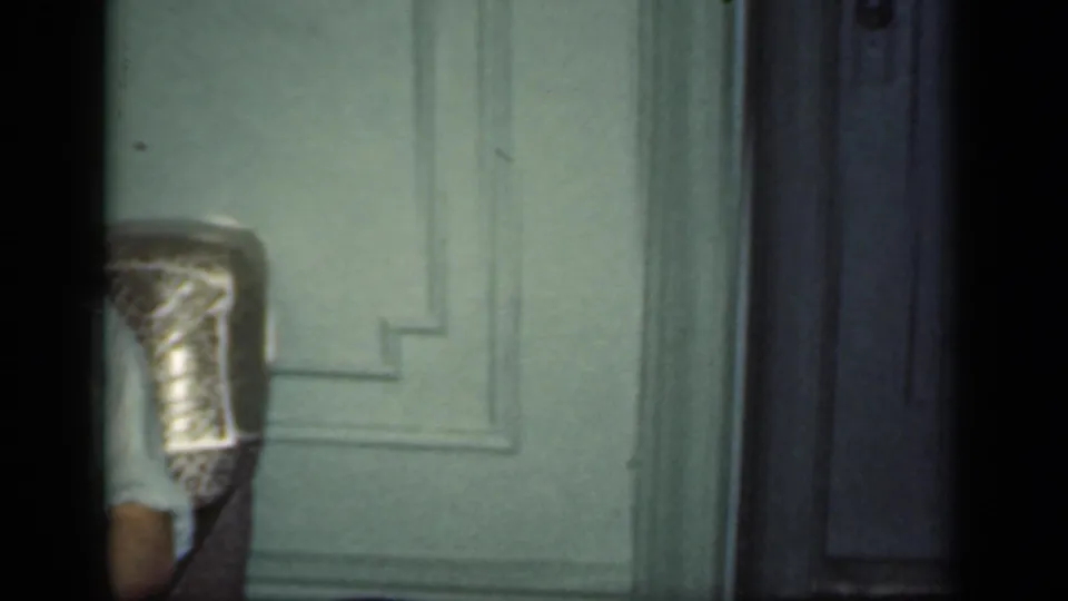 A cropped view of the armchair, pale paneled door, and wall shows the home interior as the camera moves away from the seated man.