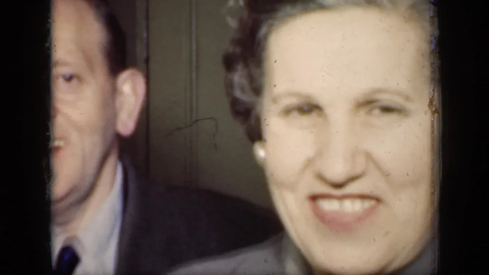 1949 New York home-movie frame showing a smiling woman close to camera with a suited man partly visible beside her in a private interior.