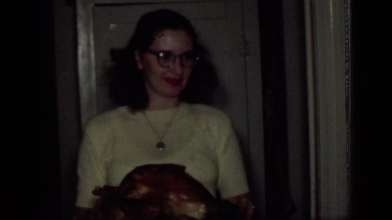 1949 New York home-movie frame of a dark-haired woman with glasses carrying a roasted chicken in a private interior doorway.