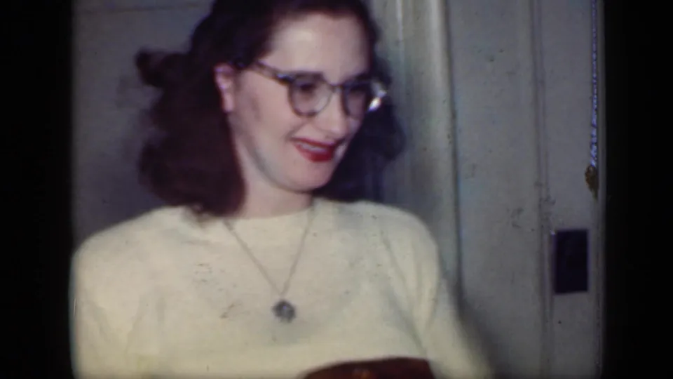1949 New York home-movie frame showing a smiling woman with glasses and pendant necklace holding a roasted chicken in a private room.