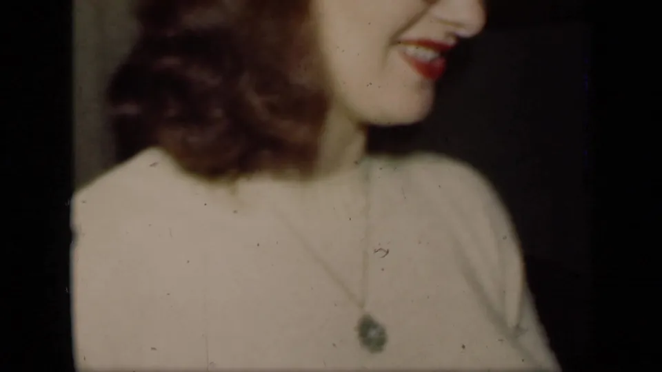 1949 New York home-movie frame with a close side view of a smiling woman in a light sweater and pendant necklace against a dark interior.