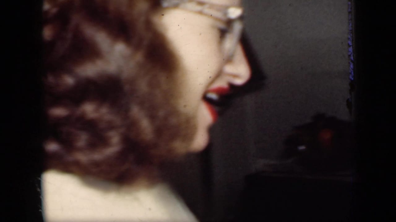 Woman with glasses smiles in profile near a dining table in a dim interior.