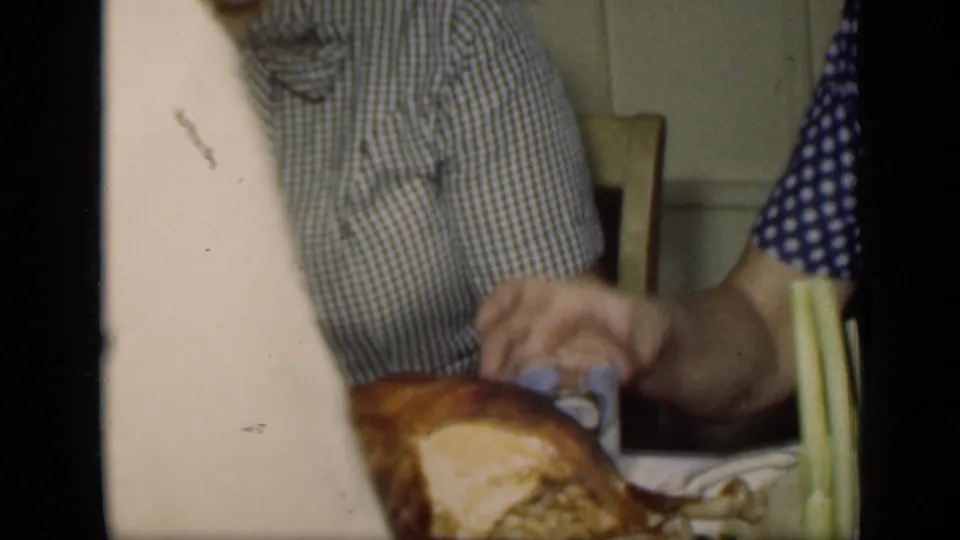 A woman in a checkered dress helps place a roasted turkey on a platter during the New York home dinner scene.