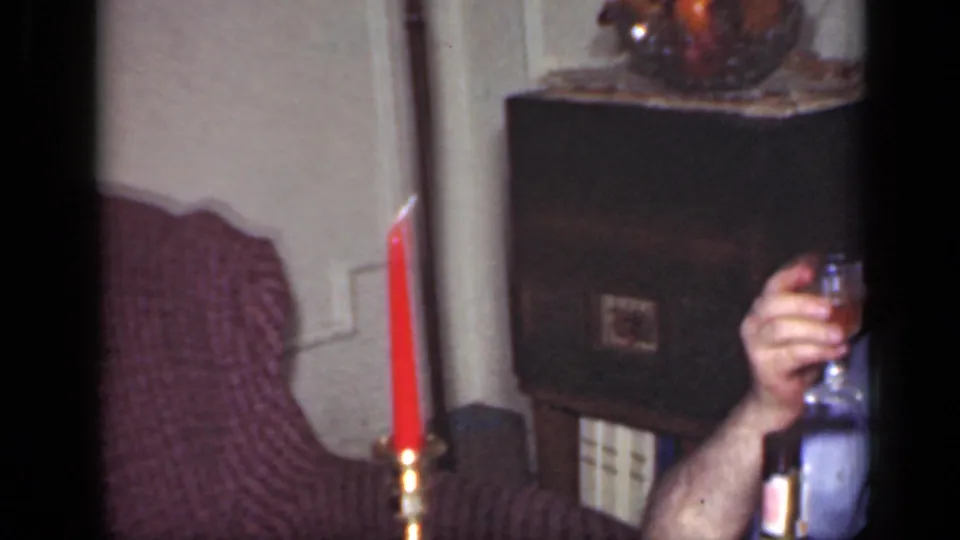 A partially framed adult sits beside a holiday table with a red candle, bottle, cabinet, and glassware in a private interior.