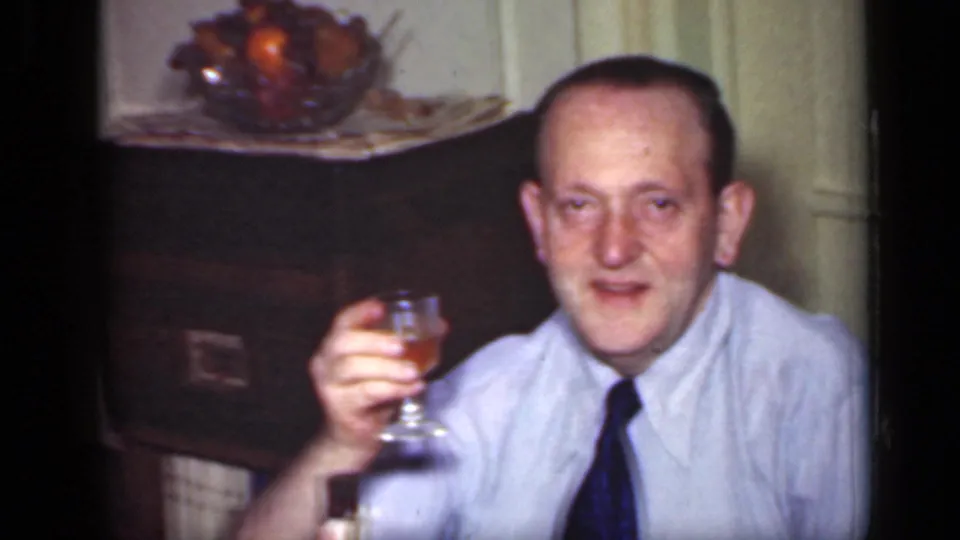 1949 New York home-movie frame of a seated man in a light shirt and dark tie raising a wine glass during dinner.