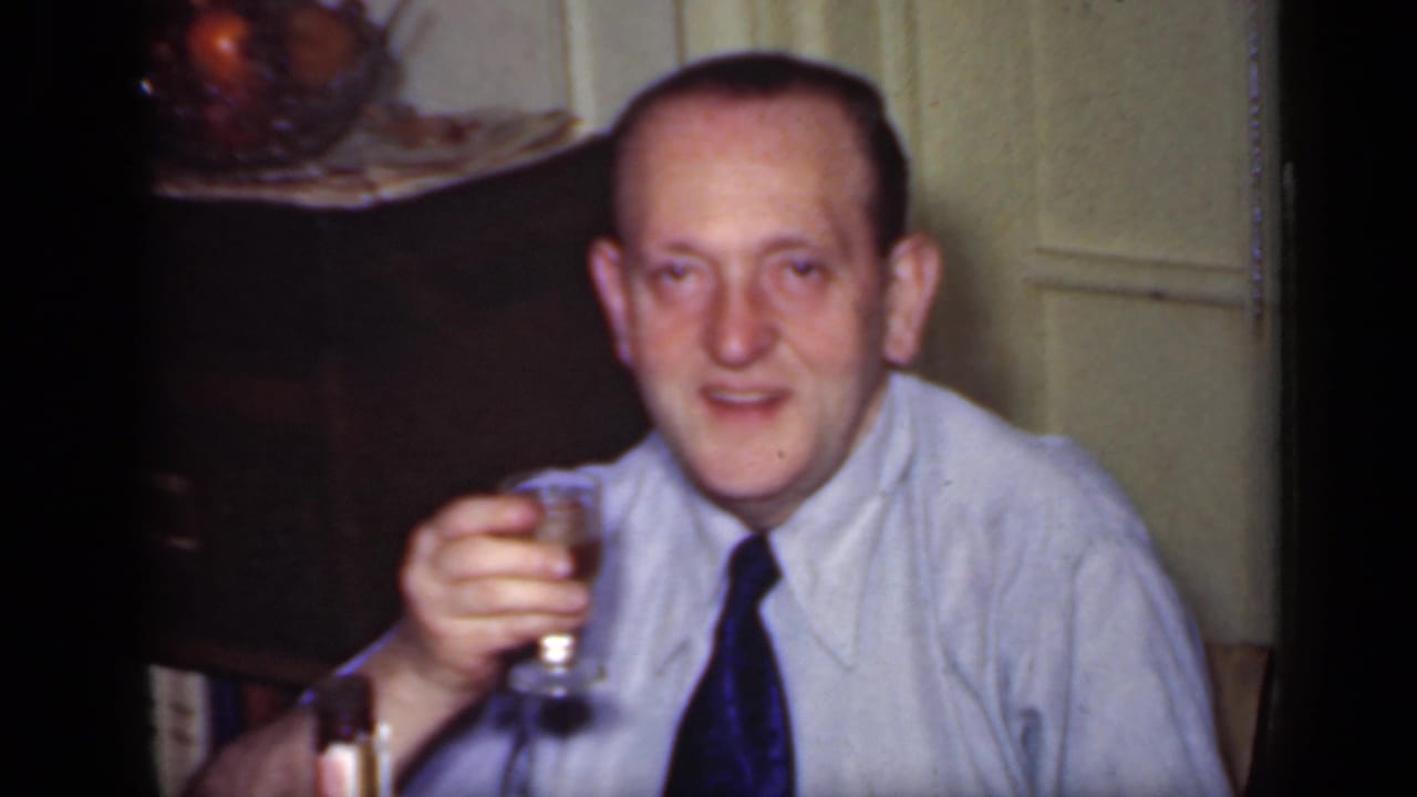 Man in a light shirt and dark tie raises a small glass while seated indoors.
