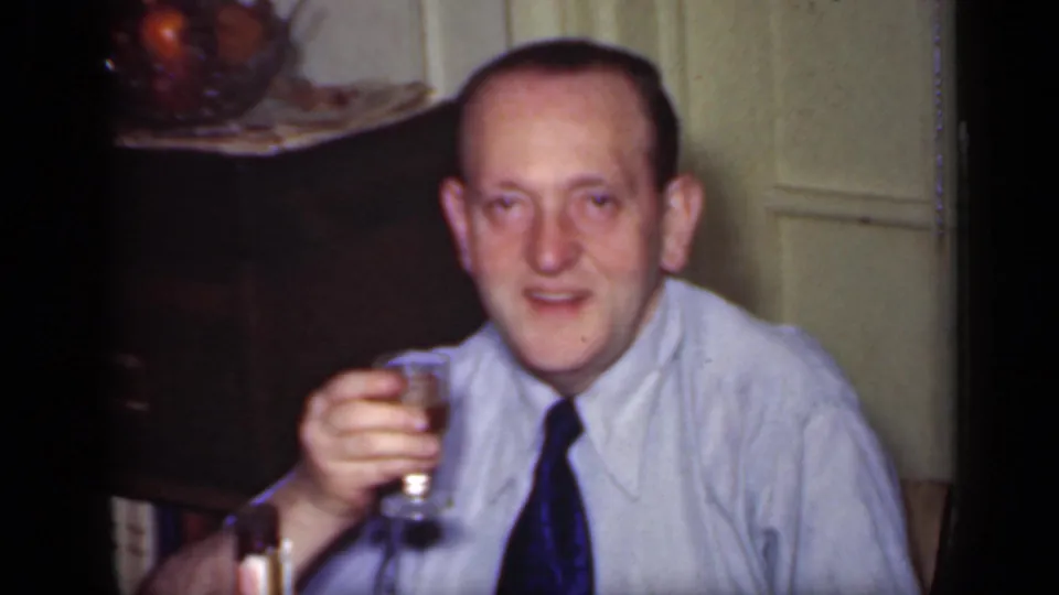 In 1949 New York home-movie footage, a seated man in a light shirt and dark tie holds up a drink toward the camera.