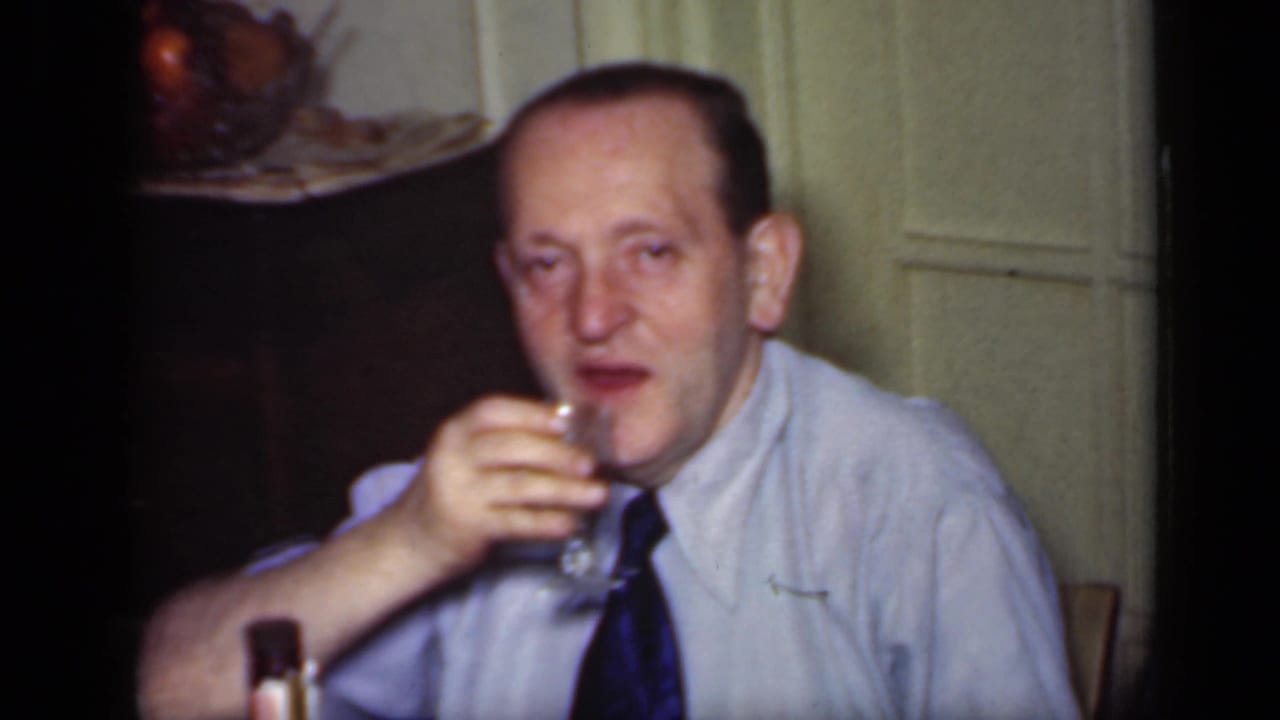 Man brings a stemmed glass to his mouth while seated beside a paneled wall.