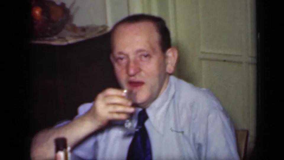 A seated man in a shirt and tie sips from a stemmed glass in an indoor 1949 New York home-movie scene.