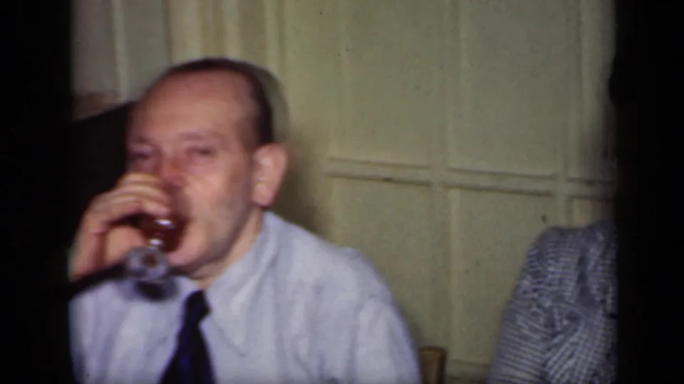 The man continues sipping from his glass, with a partial figure beside him in the softly focused indoor home-movie frame.