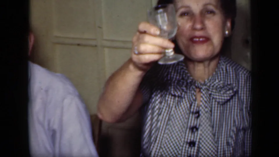 A woman in a checked blouse with a bow collar lifts a glass toward the camera in the 1949 New York indoor gathering.