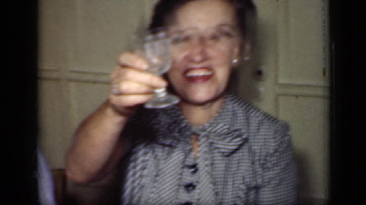 Smiling woman in a checkered blouse raises a glass toward the camera indoors.