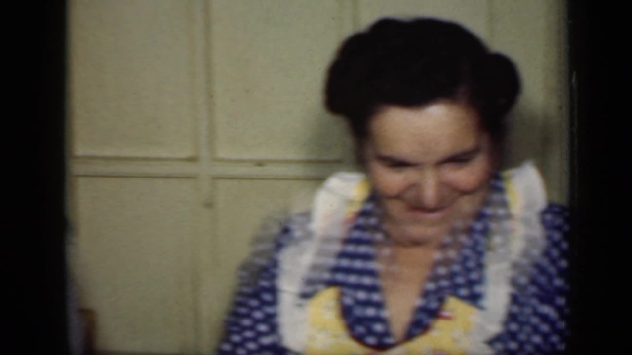 A woman in a blue polka-dot dress and yellow apron smiles with her head lowered in a 1949 New York home interior.