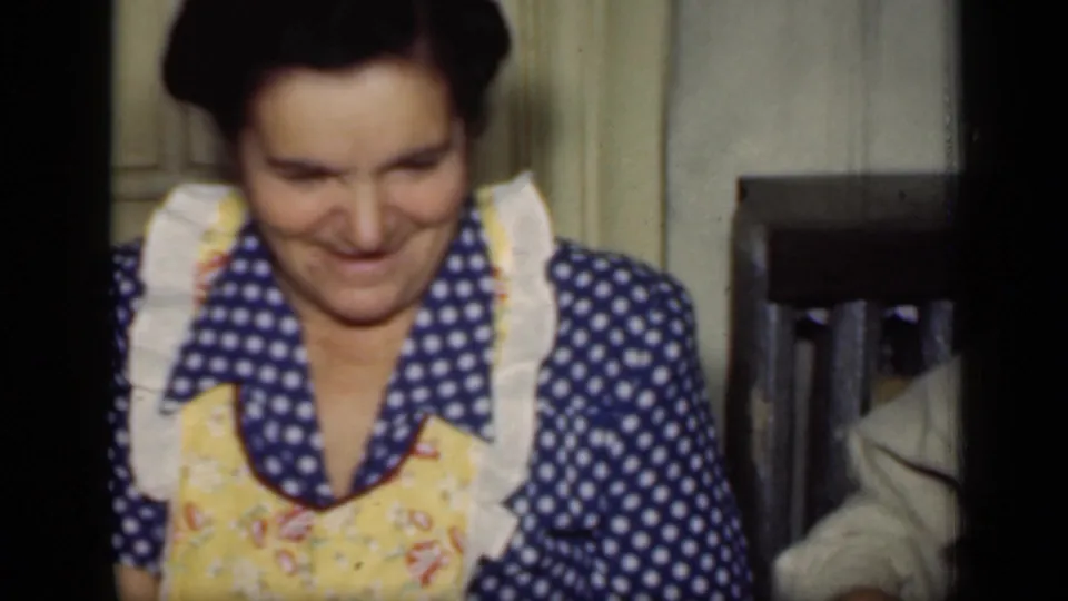 The same woman in a blue polka-dot dress and yellow apron laughs in a softly focused 1949 New York home-movie frame.