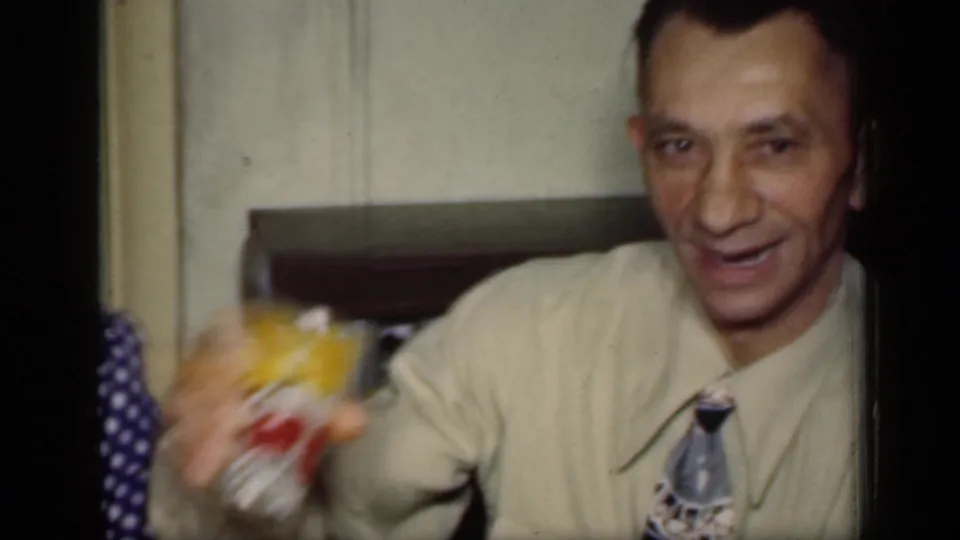 A man in a light-colored shirt and tie raises a drink toward the camera in a 1949 New York home gathering.