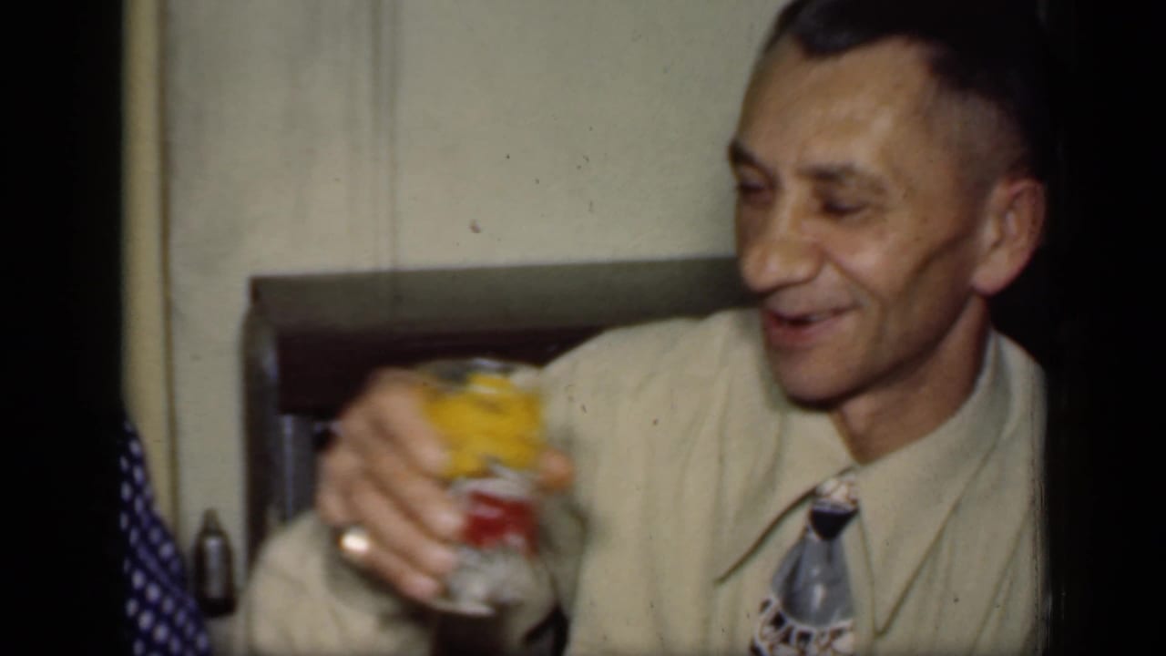 Adult man in a beige shirt and patterned tie raises a colorful glass beside an interior window.