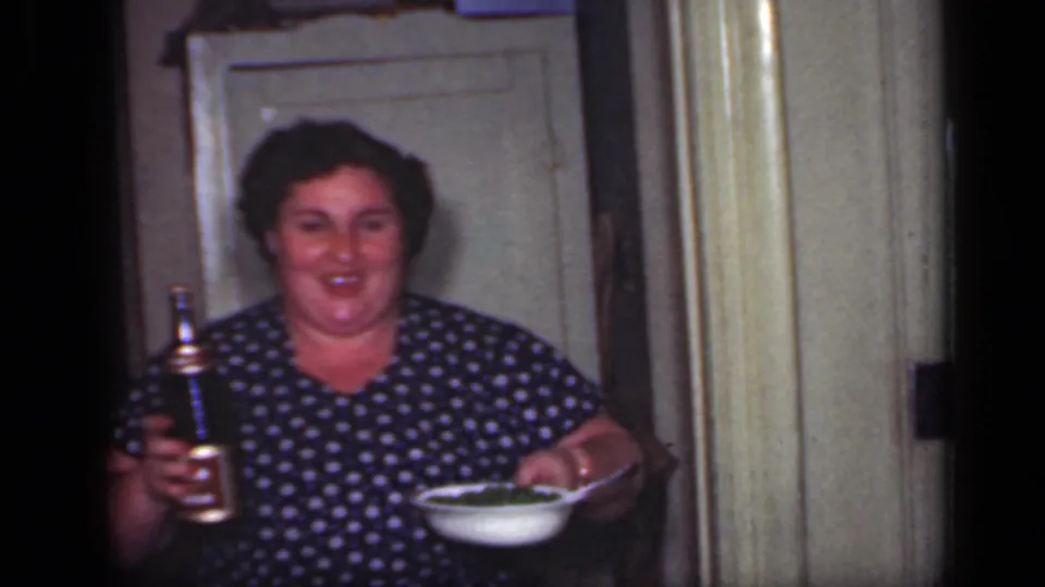 1949 New York home-movie frame of an adult woman in a blue polka dot dress holding a bottle and a bowl inside a private home.