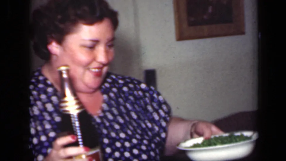 1949 New York home-movie frame of a smiling adult woman holding a bottle and a bowl of green food in a private interior.