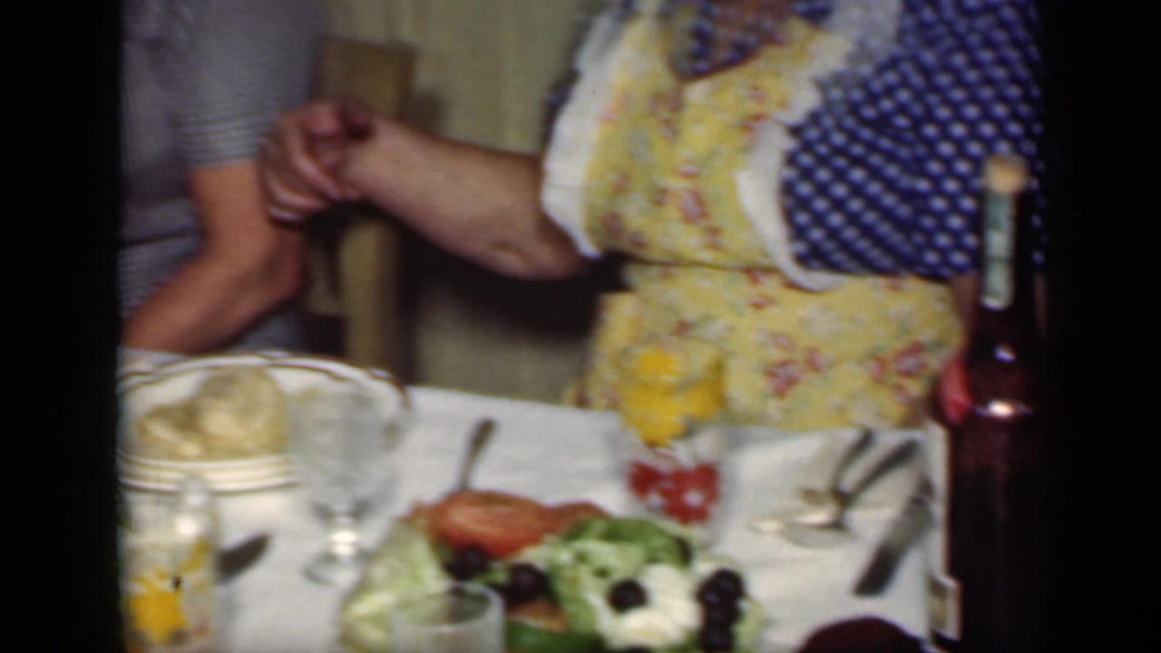 1949 New York dining-room frame with a woman in a polka dot dress and floral apron serving beside a table set with food and glassware.