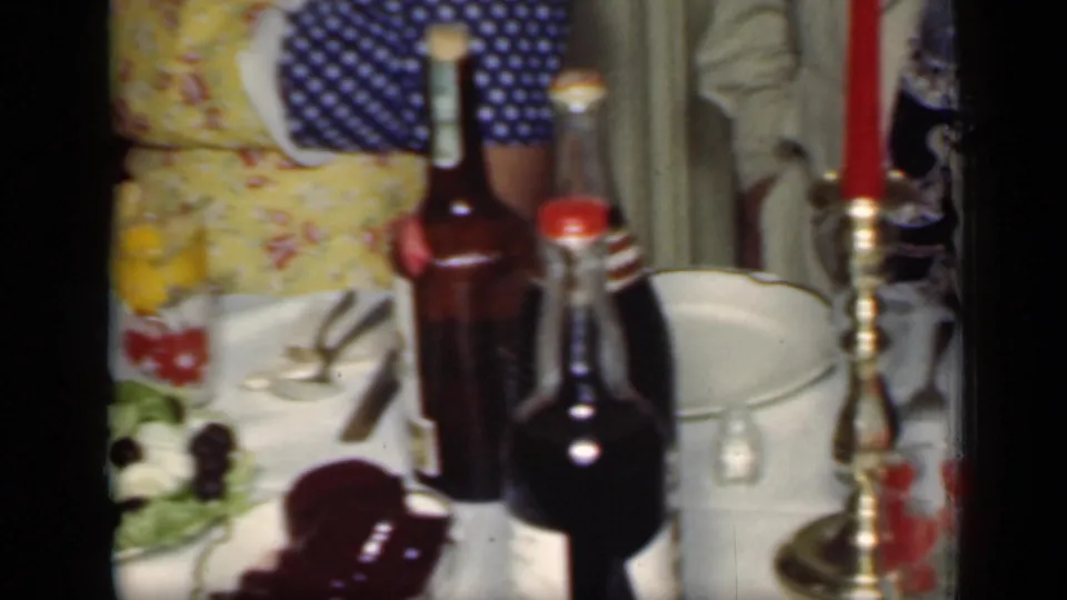 1949 New York home-movie frame showing a crowded dinner table with bottles, candlestick, plates, salad, and a woman in an apron nearby.