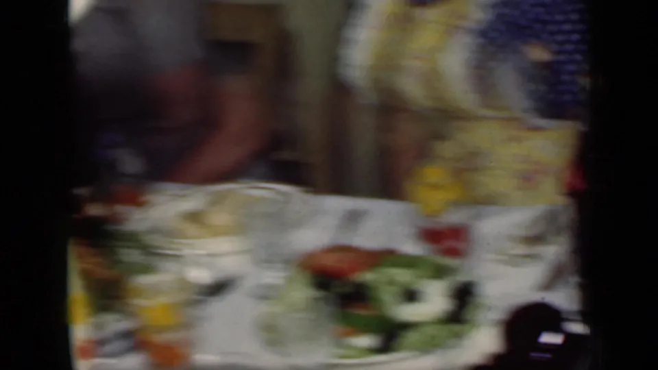 Blurred 1949 New York home-movie frame of a woman beside a dinner table with plates, salad, glassware, and serving dishes.