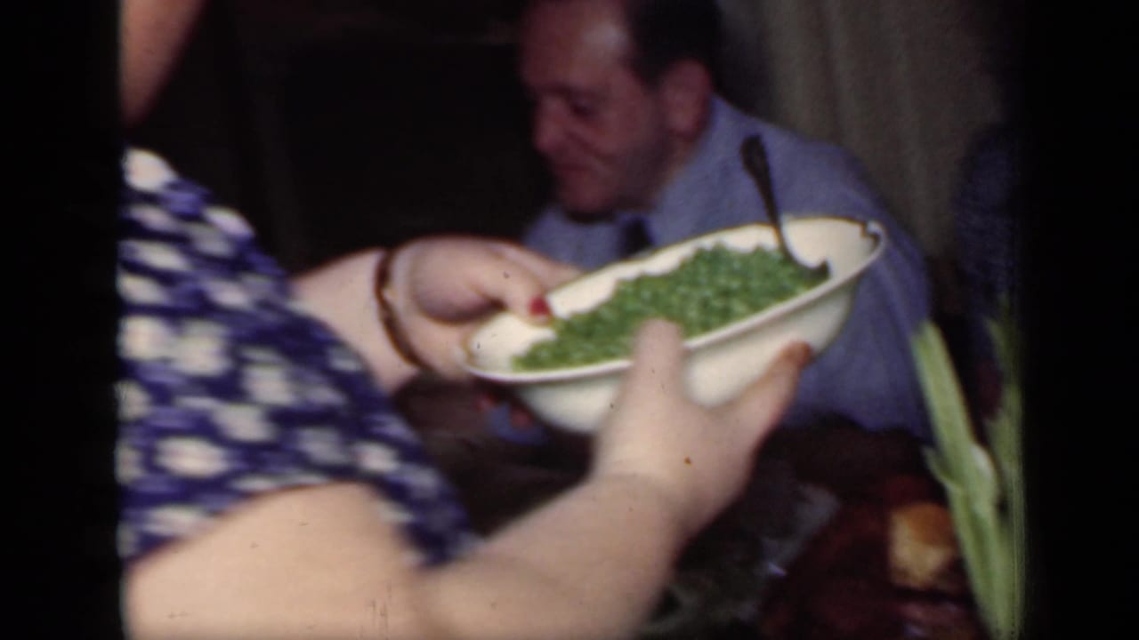 Woman holds a white bowl of green beans near a seated man at a dining table.