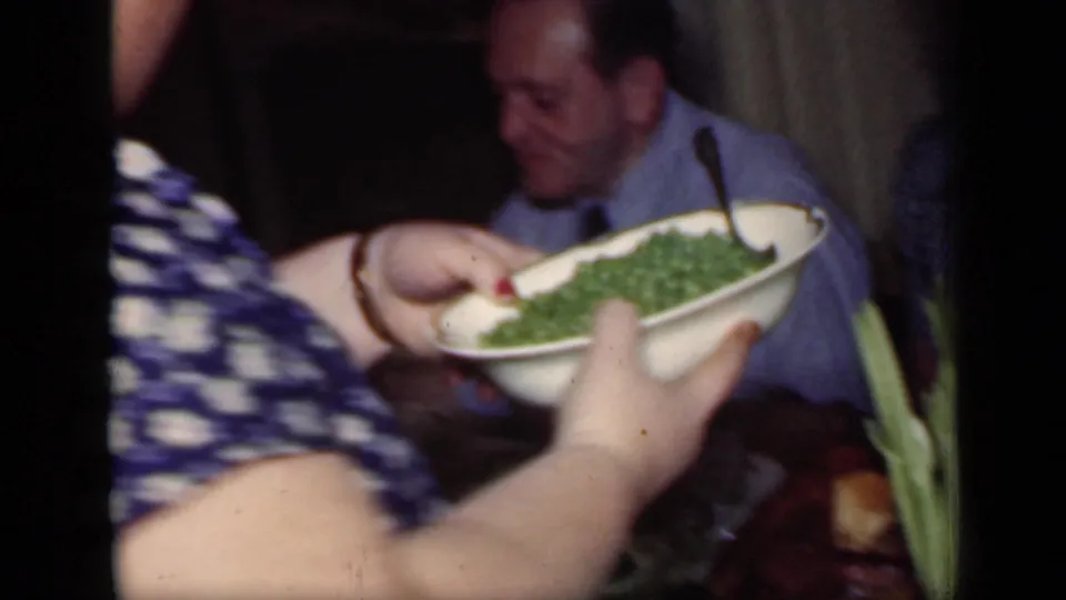 In a 1949 New York home interior, a woman in a blue polka-dot dress serves green beans beside a seated man at dinner.