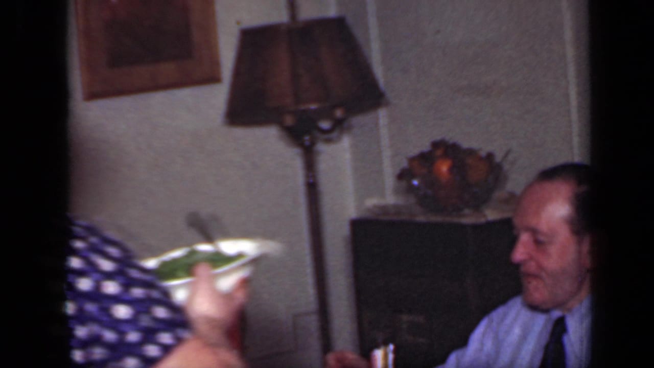 Woman passes a bowl at a dinner table while a seated man looks on near a lamp and fruit bowl.