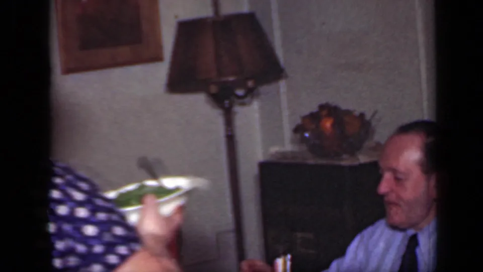 The woman continues serving food at a 1949 New York dinner table, with a seated man, table lamp, and fruit bowl in the room.