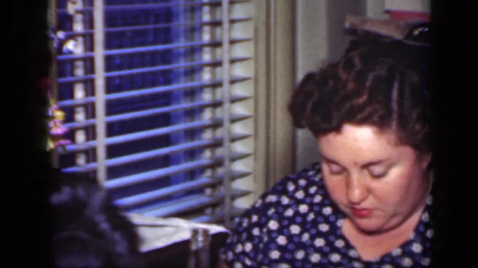 At the end of the 1949 New York dinner clip, the woman looks down toward her meal beside venetian blinds in a private home.