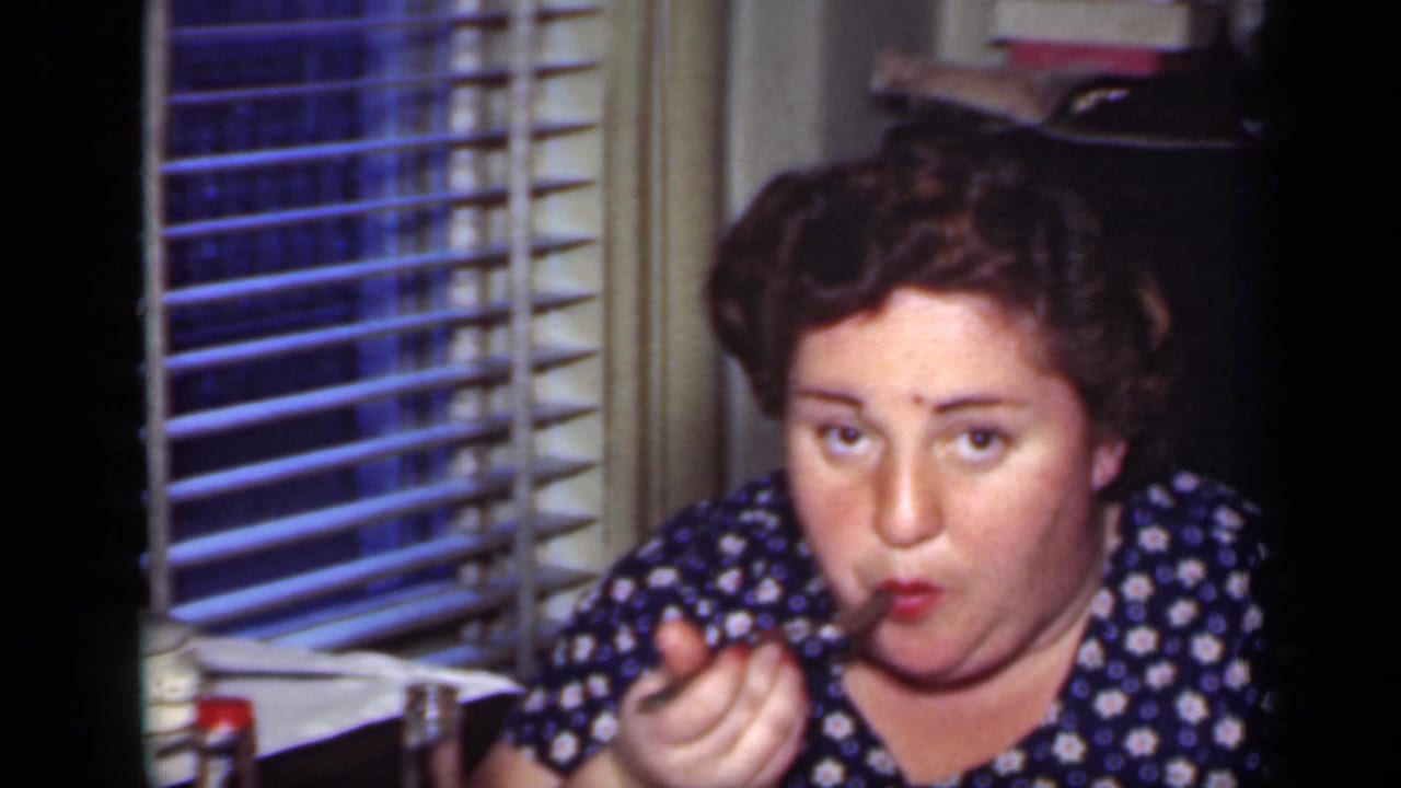 Adult woman in a blue floral dress eating from a fork indoors beside window blinds.