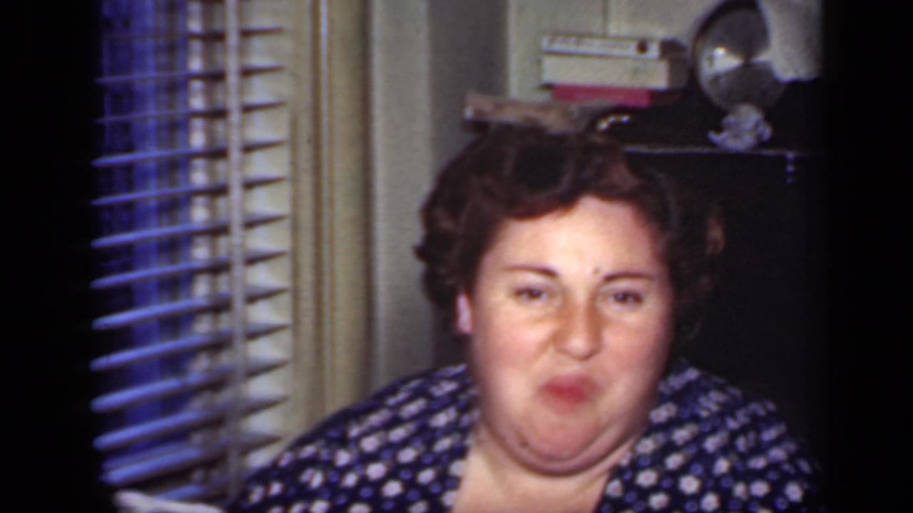 Adult woman facing the camera while chewing in a home interior with blinds behind her.