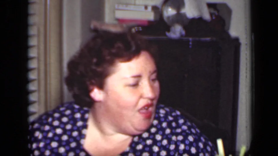 In the home-movie frame, the woman turns left with her mouth slightly open, seated in front of a dark cabinet with blinds at the side.