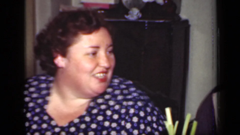 Adult woman in a blue floral dress smiles toward someone off camera, with a dark cabinet, books, and a small fan in the background.