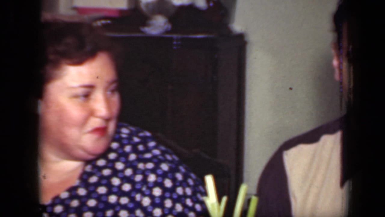 Woman in a blue polka dot dress seated indoors, looking toward someone off frame.