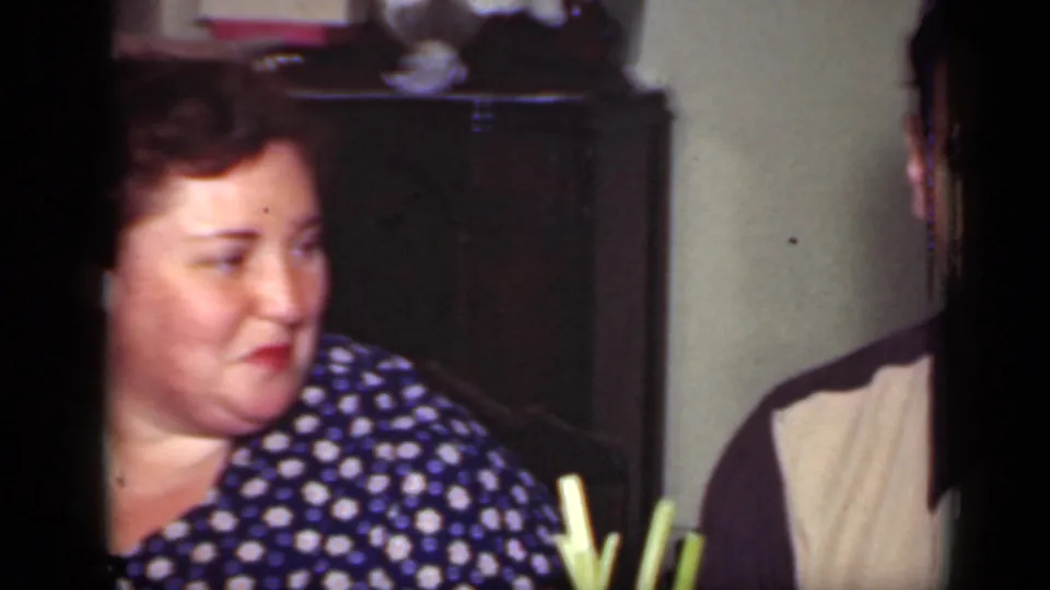 In a 1949 New York home-movie frame, a woman in a blue polka dot dress sits indoors and looks toward another person.