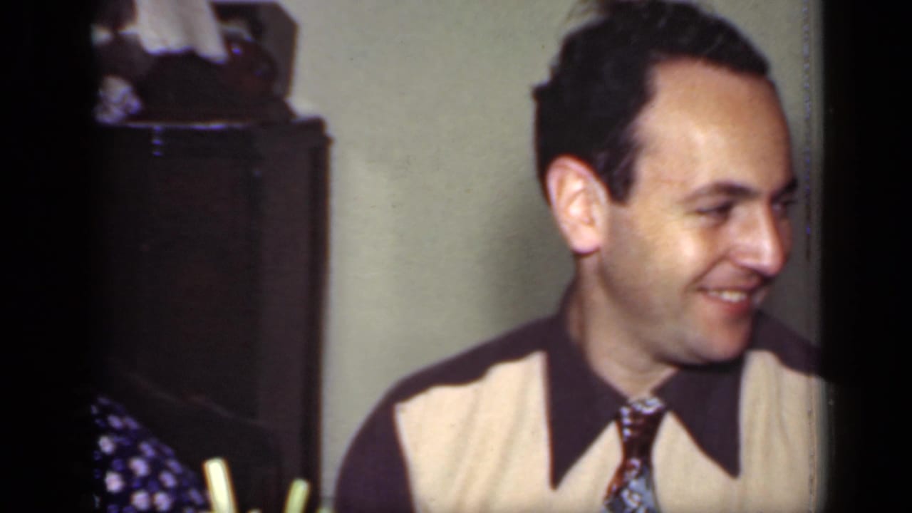A smiling man in a beige and dark sweater vest with a patterned tie sits in a New York interior from the 1949 clip.