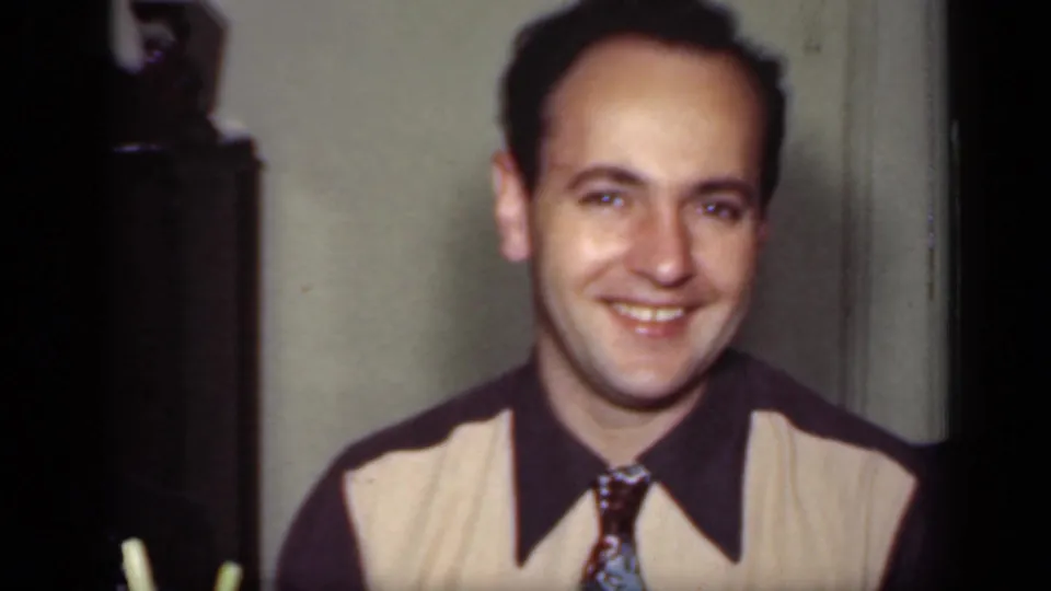 A man in mid-century clothing faces the camera and smiles inside a private New York room in the 1949 home movie.