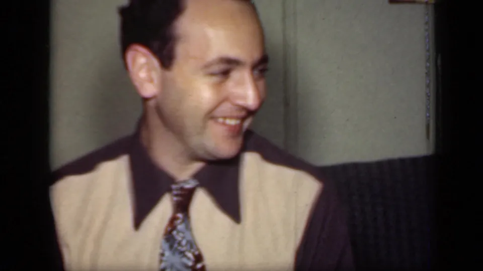 The man smiles and looks to the side, with another seated figure partly visible in the 1949 New York home interior.