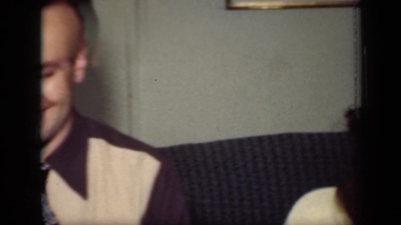 Man partly framed at left, seated beside a woman on a dark couch indoors.