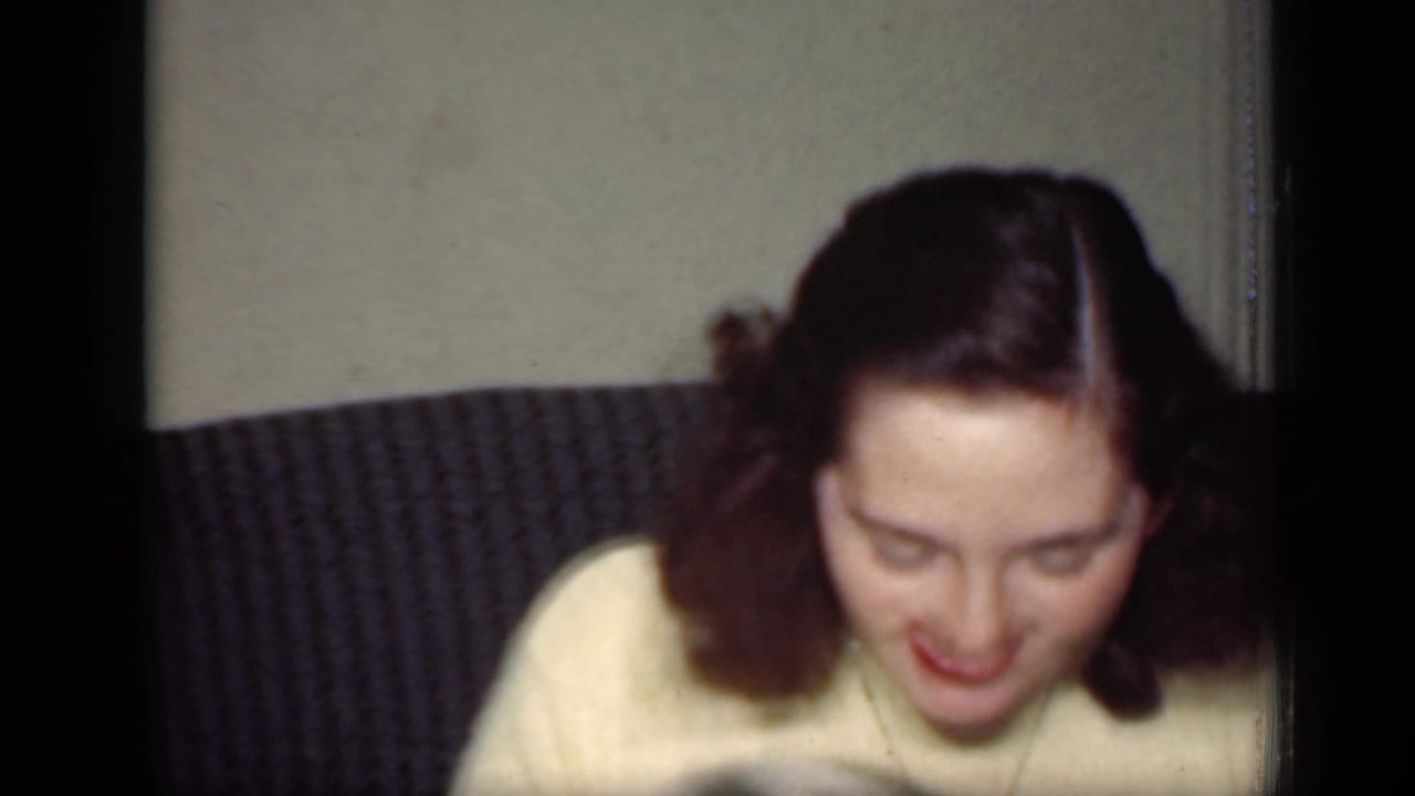 A woman with styled dark hair and red lipstick looks down from a dark couch in the 1949 New York home interior.