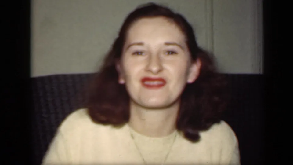 The woman smiles toward the camera in a late-1940s New York home-movie frame, seated before a dark textured couch.