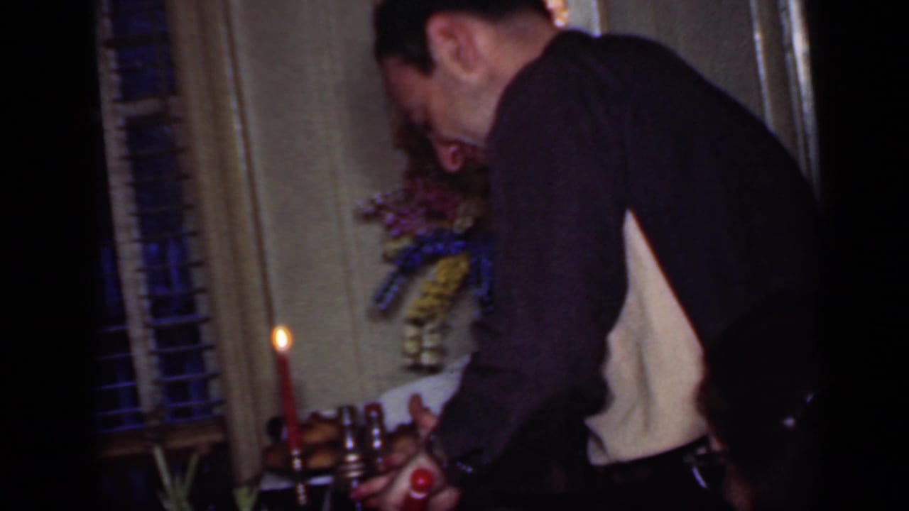 Man in a dark sweater leans over a candlelit dining table in a New York home interior, with blinds and a floral decoration behind him.