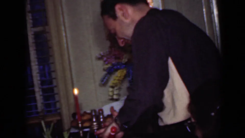 Man in a dark sweater leans over a candlelit dining table in a New York home interior, with blinds and a floral decoration behind him.